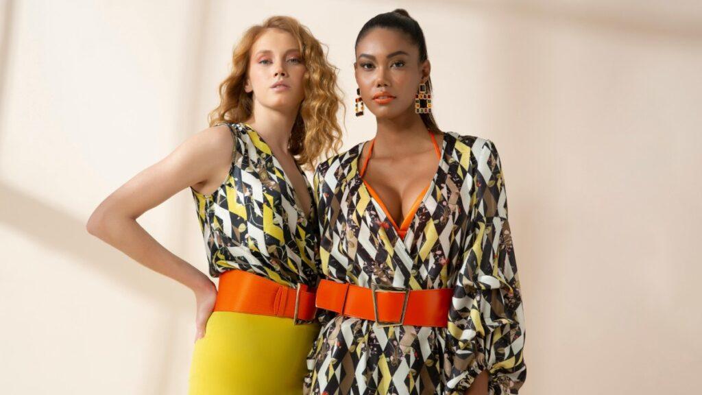 Two stylish women posing in colorful dresses with orange belts